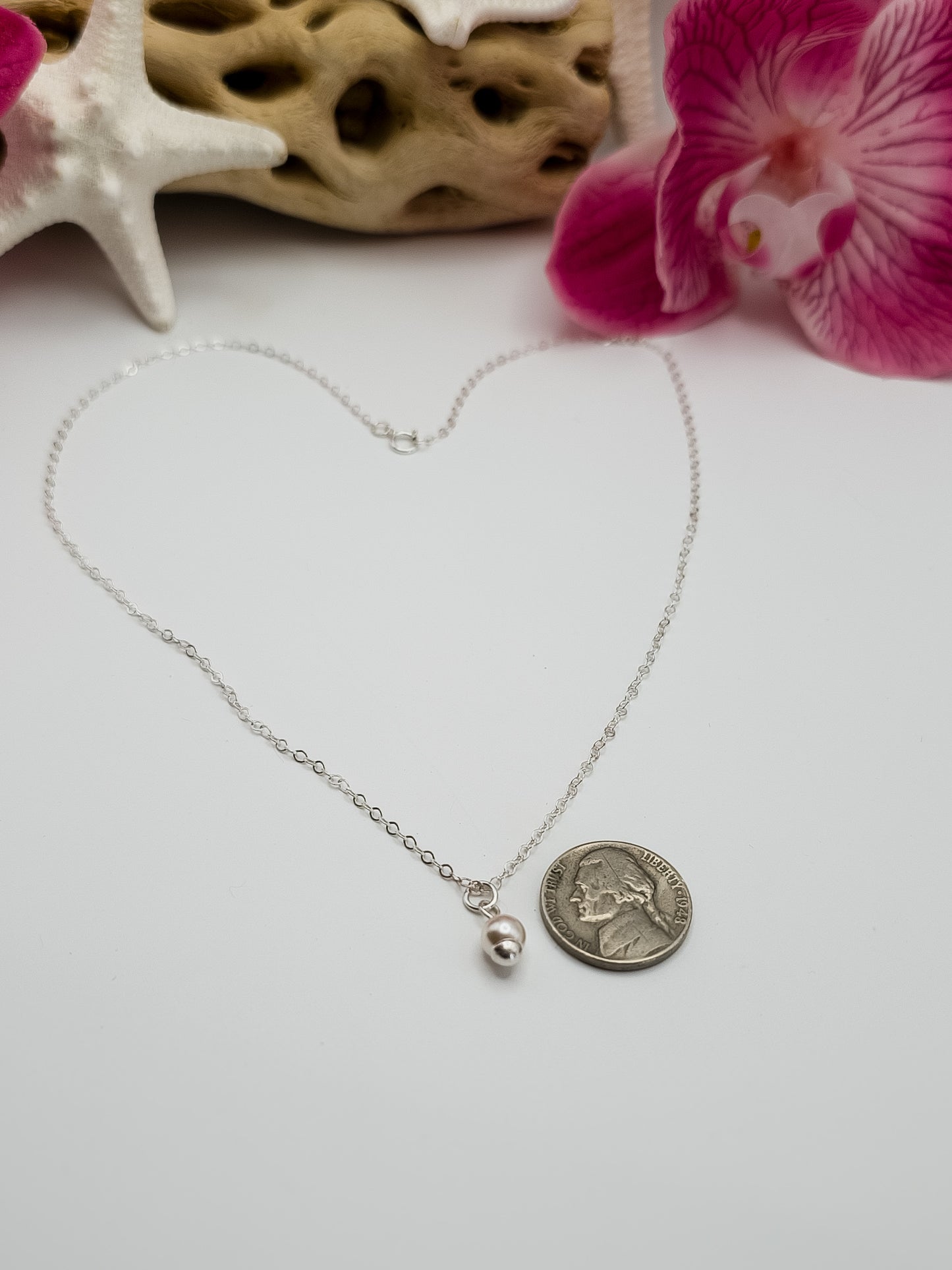 Dainty sterling silver single pearl necklace laying on a white background with a nickel next to the pearl to show the size of the pearl. Beach decor in the background for staging.