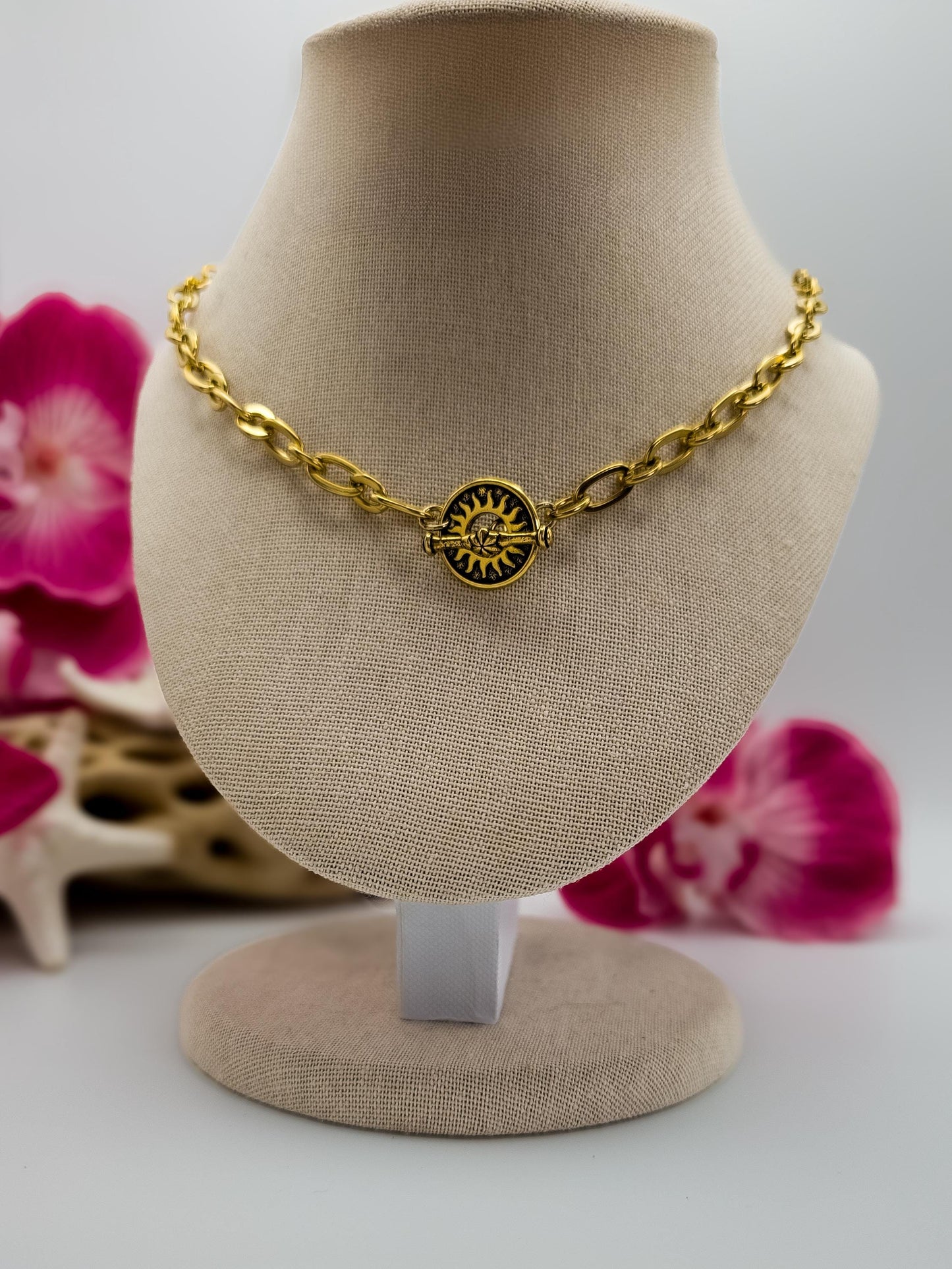 A gold-toned oval link chain necklace is displayed on a light brown jewelry bust. The necklace features a central, round toggle clasp as a pendant with a stylized sun design in gold and black. The sun has radiating lines and a central circle with smaller details.