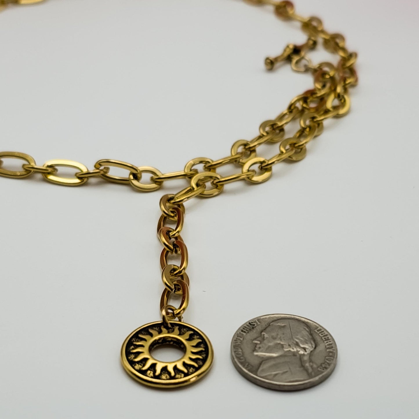 Close-up of a gold sun pendant attached to an oval link chain, positioned beside a US nickel to show its dimensions. The pendant has intricate radiating lines.