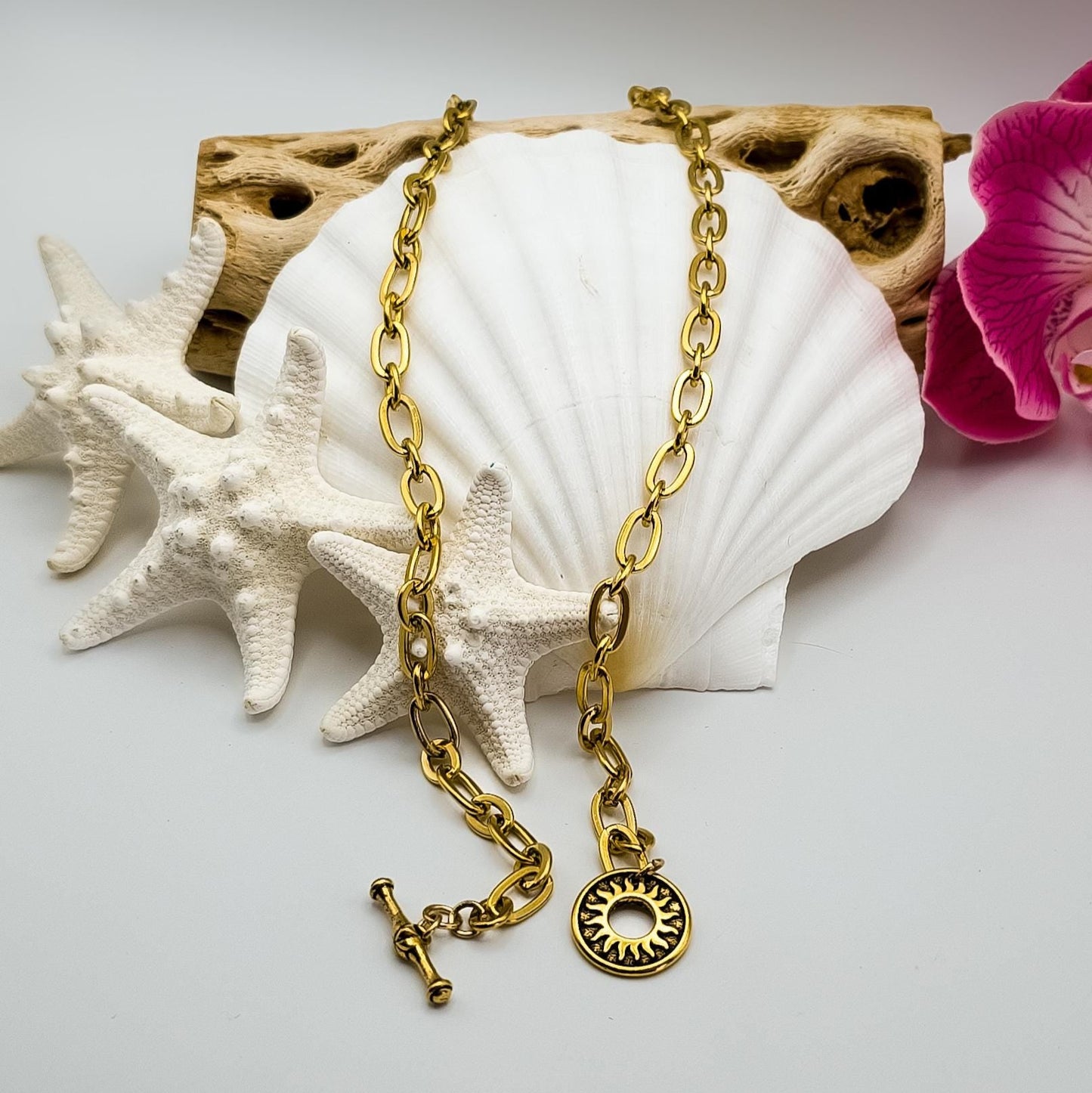 A chunky gold-toned oval link chain necklace is displayed on a white seashell, surrounded by white starfish and driftwood. A pink orchid is visible on the right.