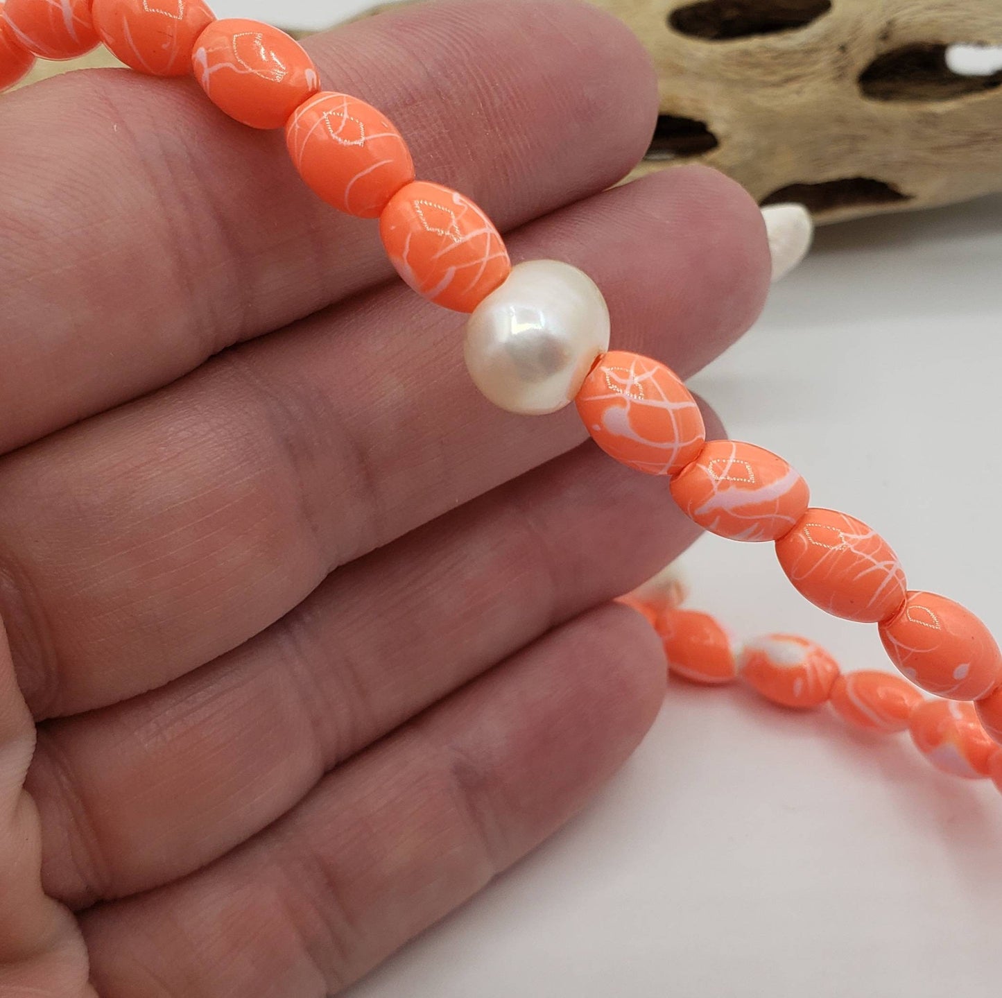 A hand gently holds a stretchy orange beaded anklet, showcasing a central freshwater pearl and the size of the vibrant orange beads.