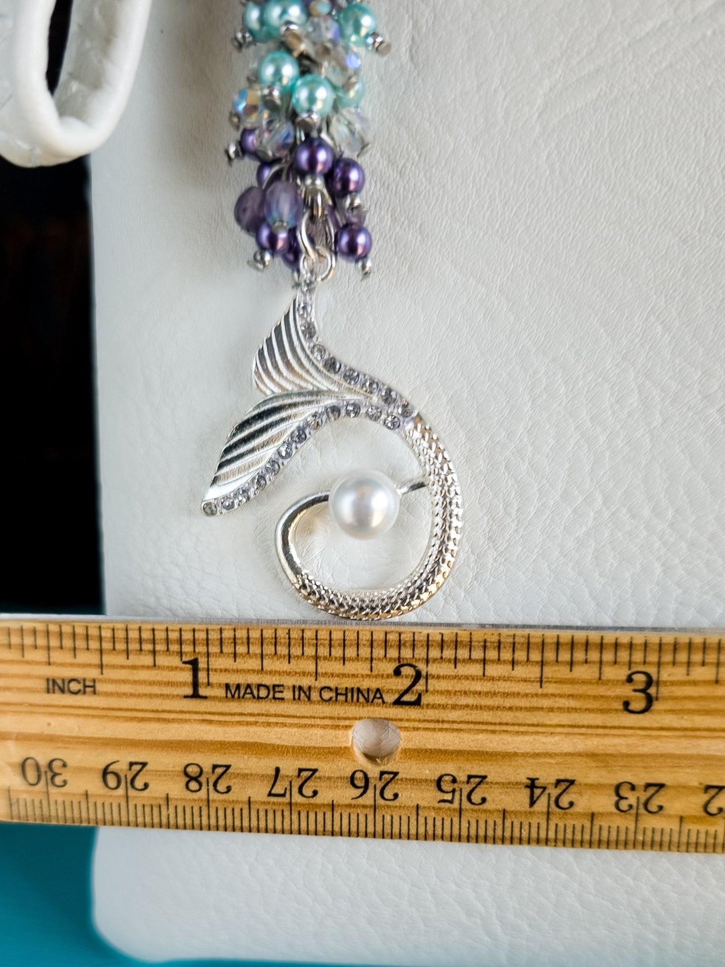 Close-up of a mermaid bag charm dangling from a purse, with a wooden ruler indicating a width of about one inch across the silver mermaid tail.