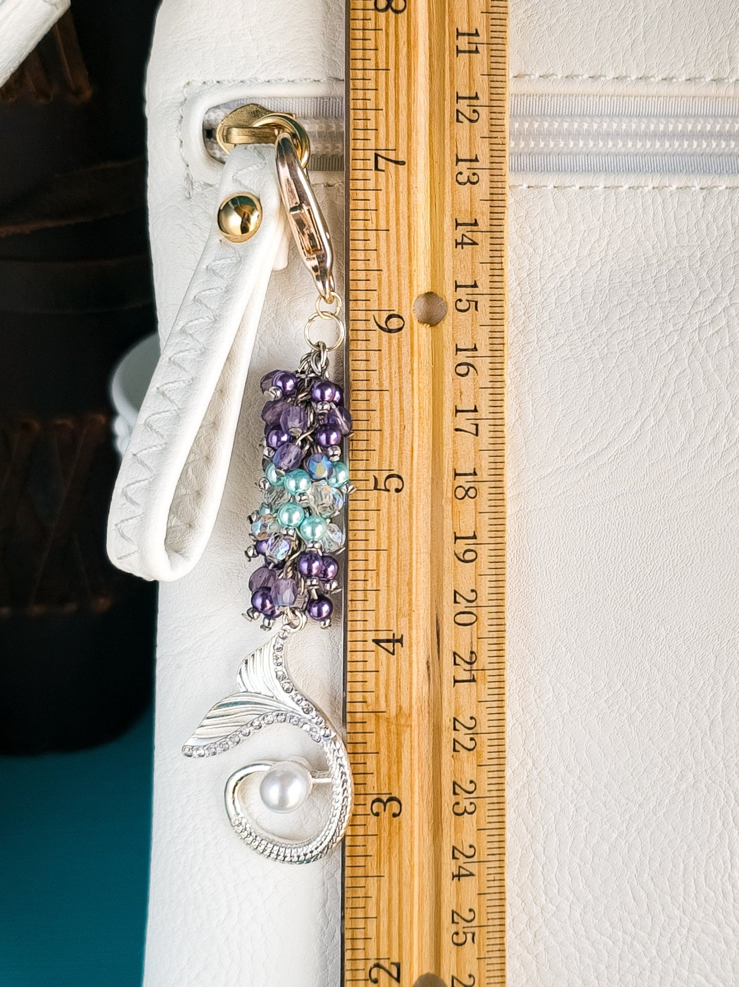 A mermaid bag charm, approximately 4.5 inches long as indicated by a wooden ruler, hangs from a white purse. The charm features purple and turquoise pearls, AB crystals, and a silver-plated mermaid tail.