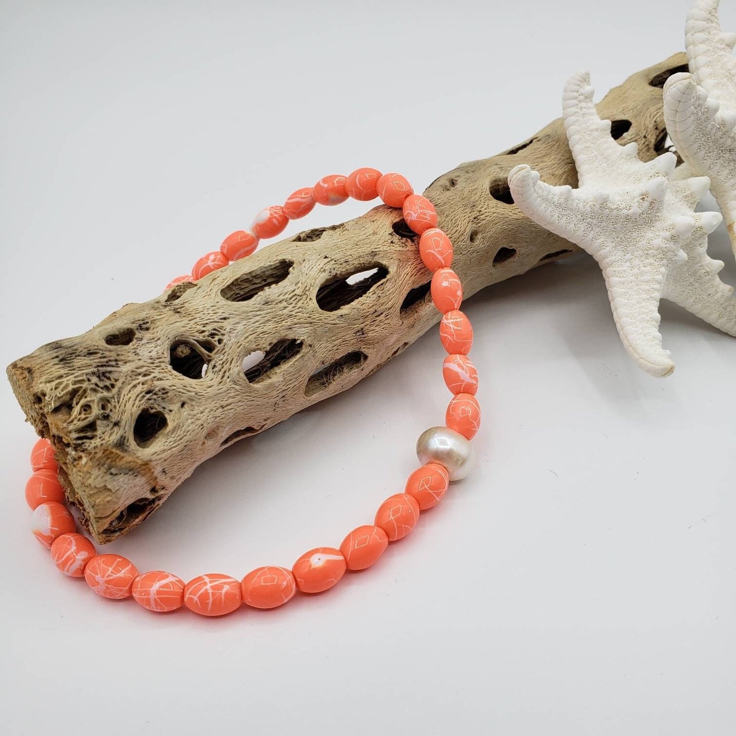 Stretchy orange beaded anklet featuring a single freshwater pearl as the focal point. The anklet rests across a collection of various seashells and weathered driftwood, suggesting a beachy setting.