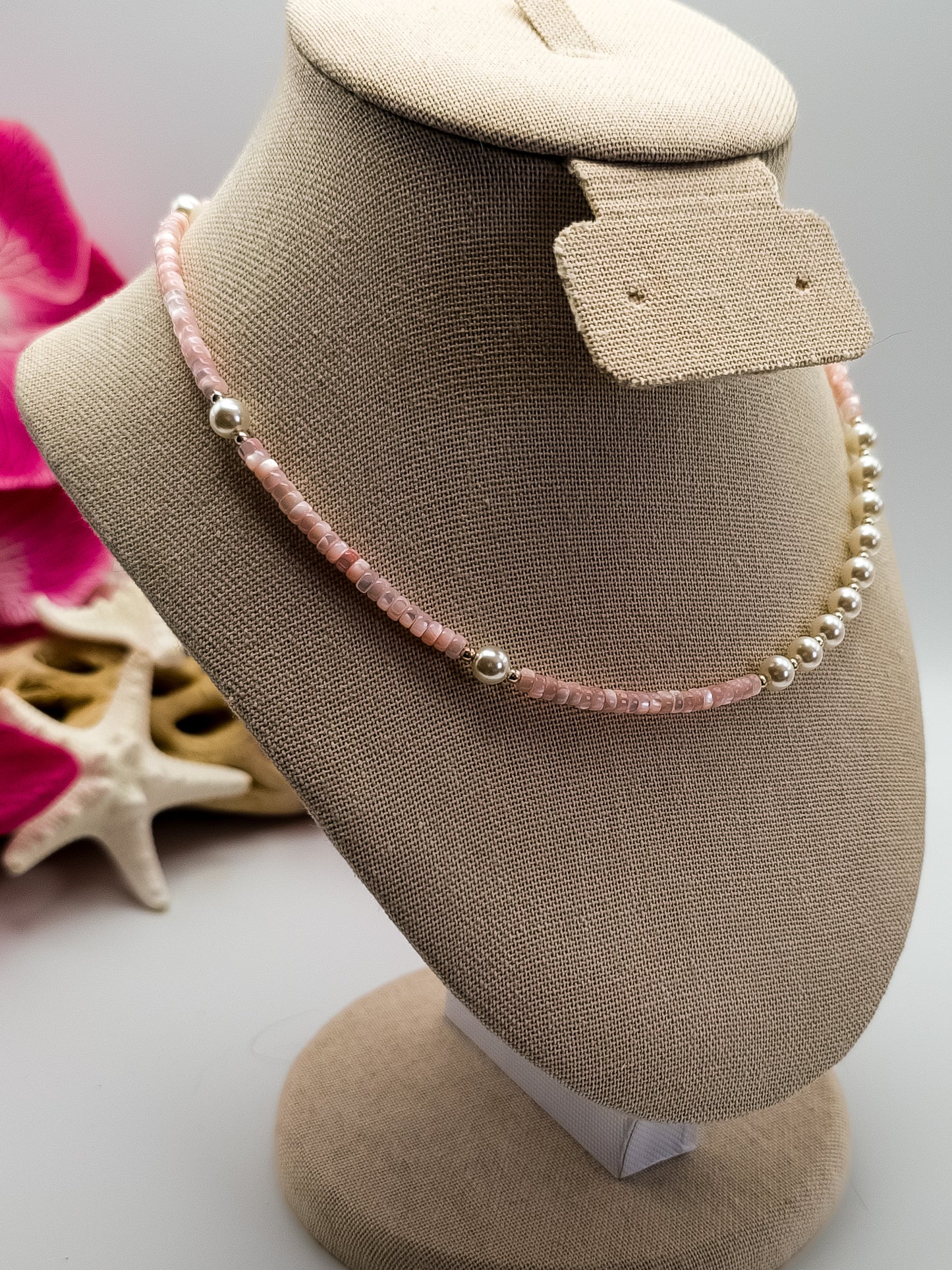 Light pink Mother of Pearl heishi beads with small silver plated beads and white glass pearls handmade choker laying on a tan neck display with flowers and driftwood in the background for staging.