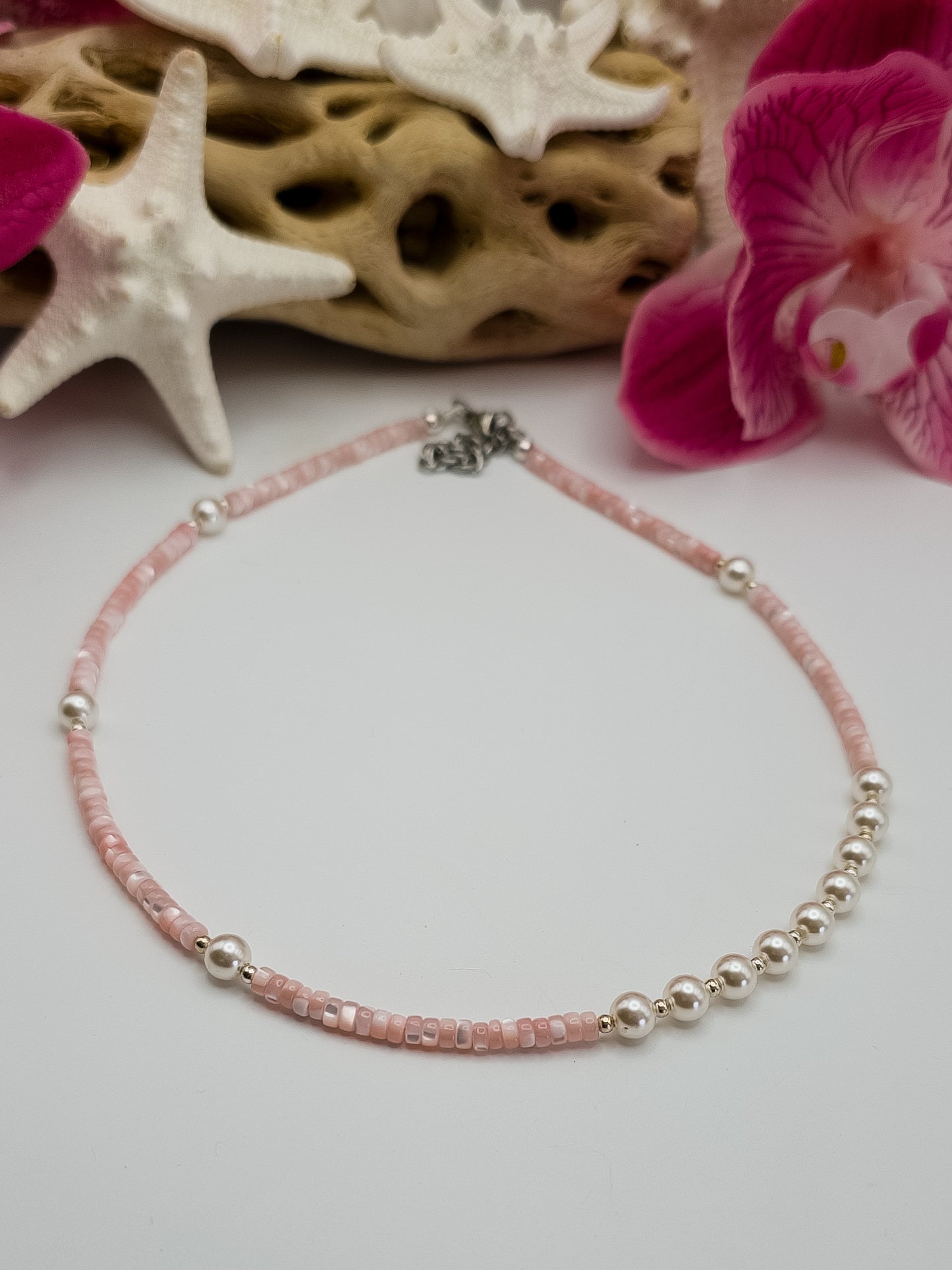 Light pink Mother of Pearl heishi beads with small silver plated beads and white glass pearls handmade choker laying on a white surface with starfish, flowers and driftwood in the background for staging.