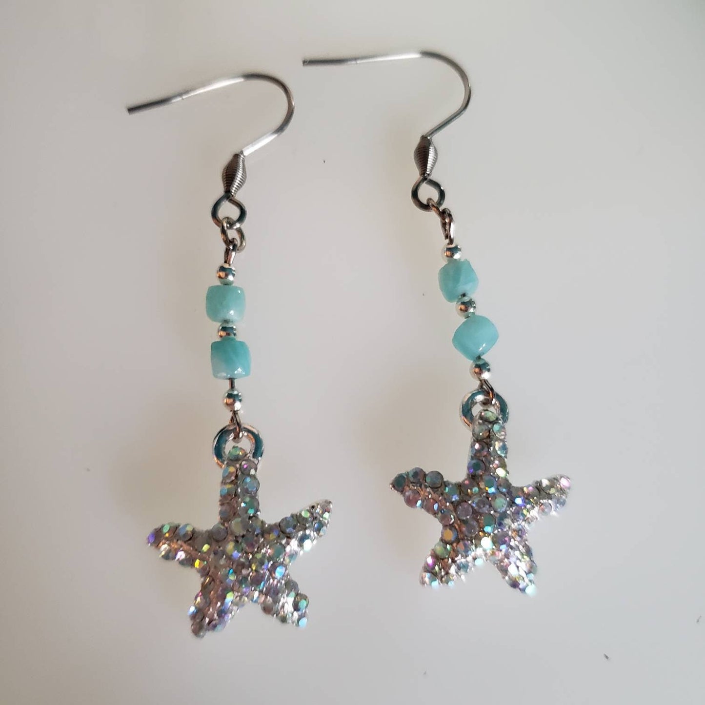 Silver starfish earrings adorned with sparkly rhinestones and blue-green amazonite beads rest on a white surface. The starfish shape and gemstone colors are clearly visible.
