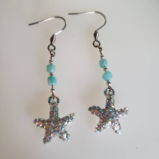 Silver starfish earrings adorned with sparkly rhinestones and blue-green amazonite beads rest on a white surface. The starfish shape and gemstone colors are clearly visible.