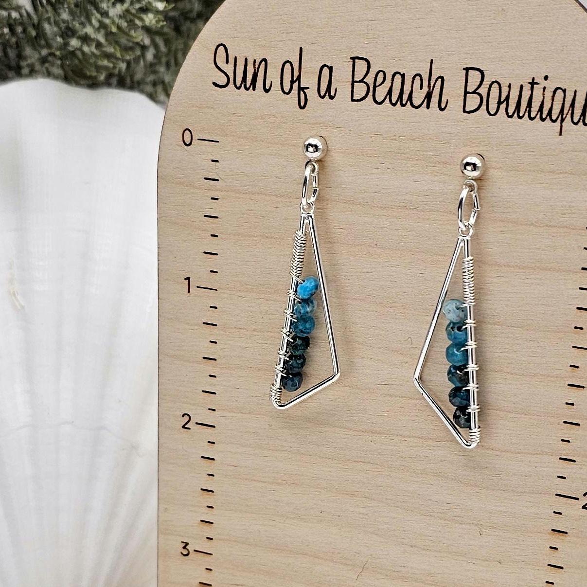 Silver triangle earrings with blue gemstone beads hang from an earring holder. The text "Sun of a beach boutique" is visible at the top of the holder. 