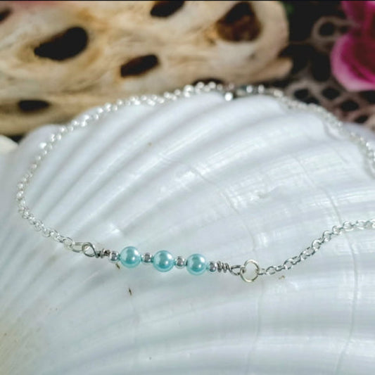 A delicate silver chain anklet with three light blue aqua glass pearls displayed on a textured white seashell.