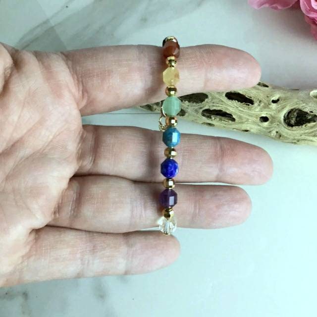 A chakra bracelet with a gold-filled chain features a sequence of gemstone beads in clear, purple, blue, light blue, green, yellow, orange, and red, representing the chakras. The bracelet is being held by fingers to show the size. There is a piece of driftwood in the background. 
