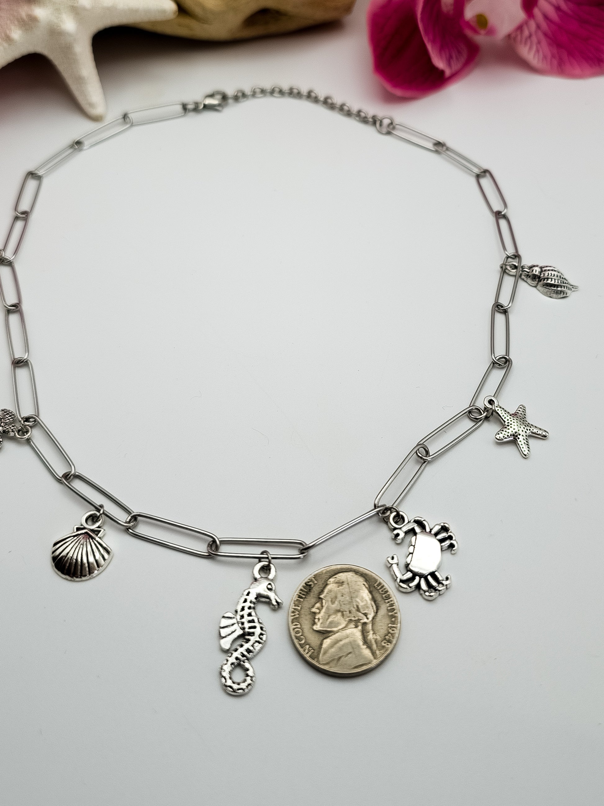 Stainless steel paperclip chain with beach charms in silver attached to the necklace. It is laying flat on a white surface. The charms are a mermaid, sea turtle, seashell, seahorse, crab, and starfish. Driftwood, fake starfish and flower in the background for staging. 
