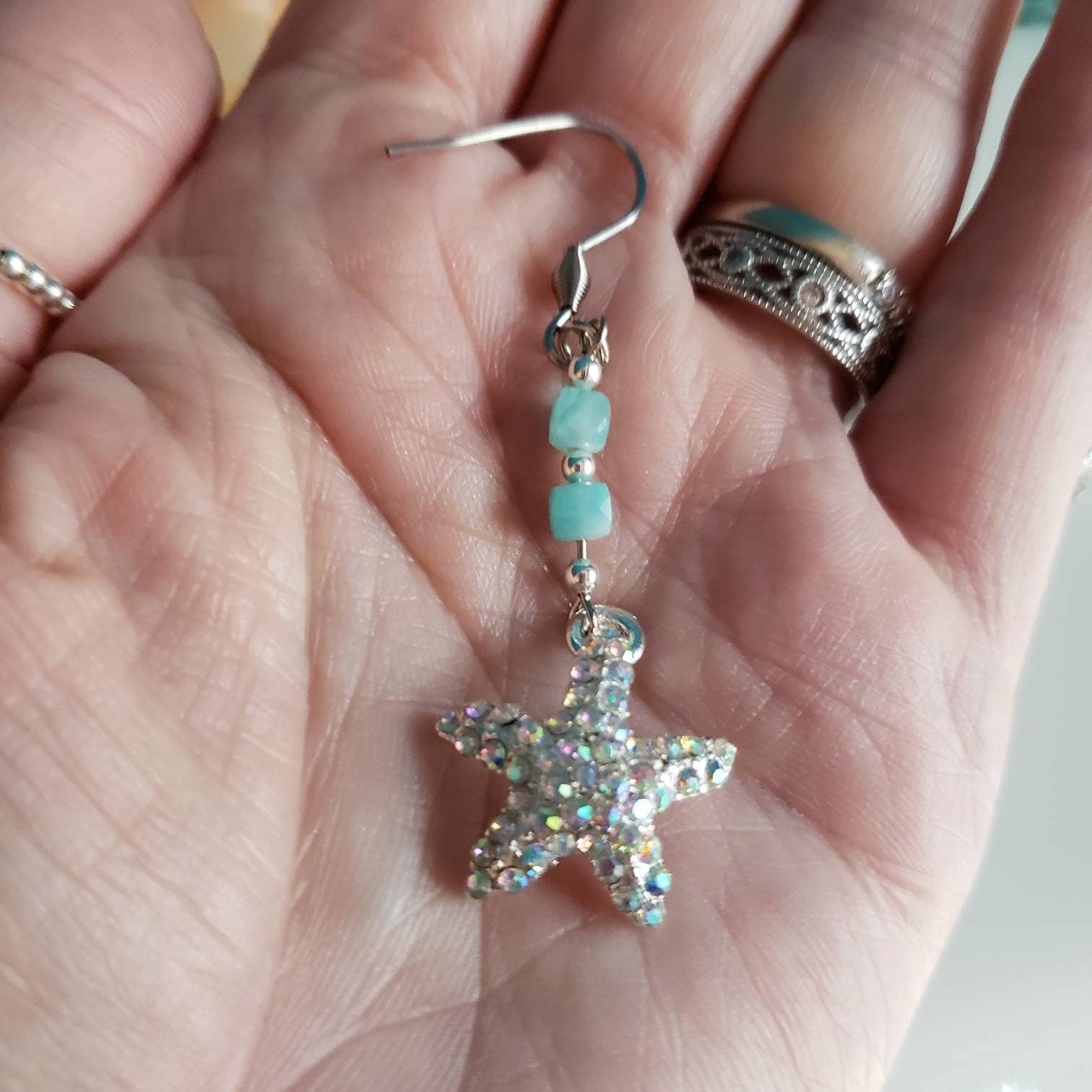 A hand gently holds a silver starfish earring, showcasing its size and the detail of the sparkly rhinestones and blue-green amazonite beads.