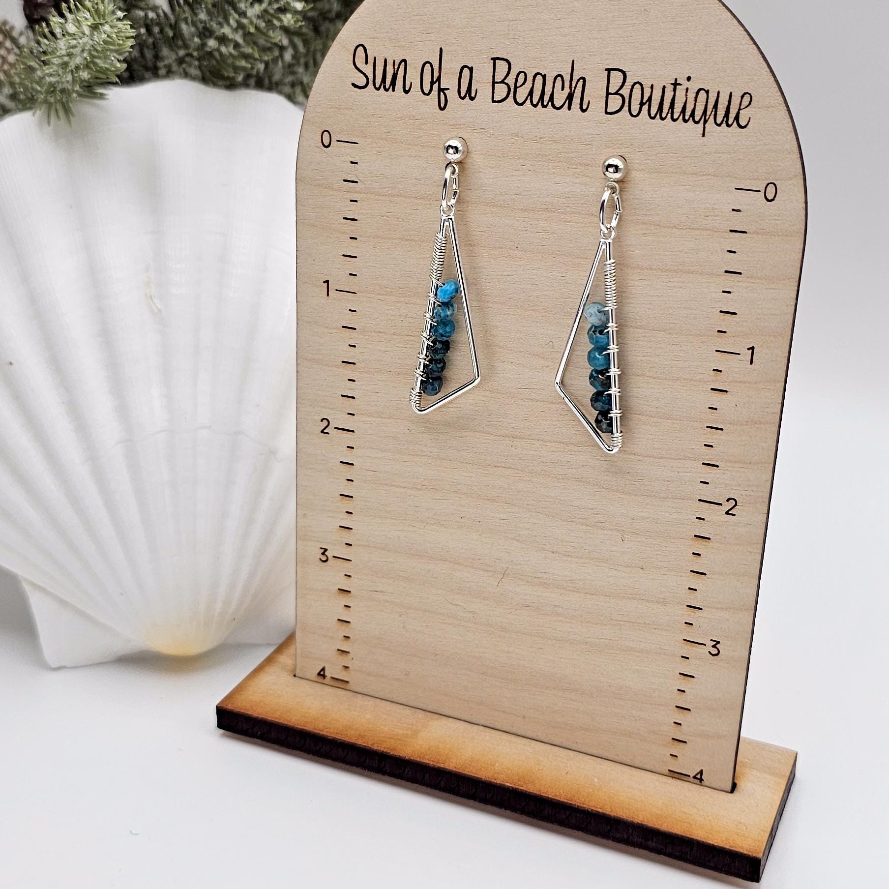 Silver triangle earrings with blue gemstone beads hang from an earring holder. The text "Sun of a beach boutique" is visible at the top of the holder. There is a seashell and some greenery in the background. 