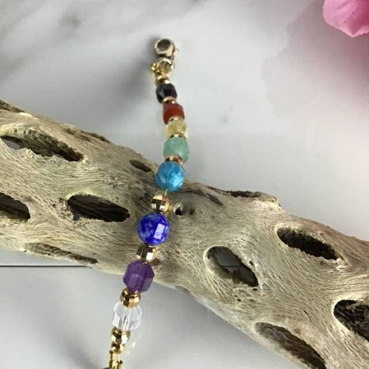 A chakra bracelet with a gold-filled chain features a sequence of gemstone beads in clear, purple, blue, light blue, green, yellow, orange, and red, representing the chakras. The bracelet rests on a piece of driftwood. 