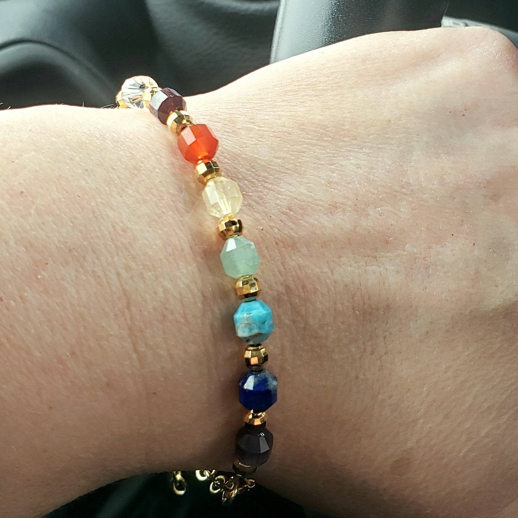 A chakra bracelet with a gold-filled chain features a sequence of gemstone beads in clear, purple, blue, light blue, green, yellow, orange, and red, representing the chakras. The bracelet is shown being worn on a wrist. 