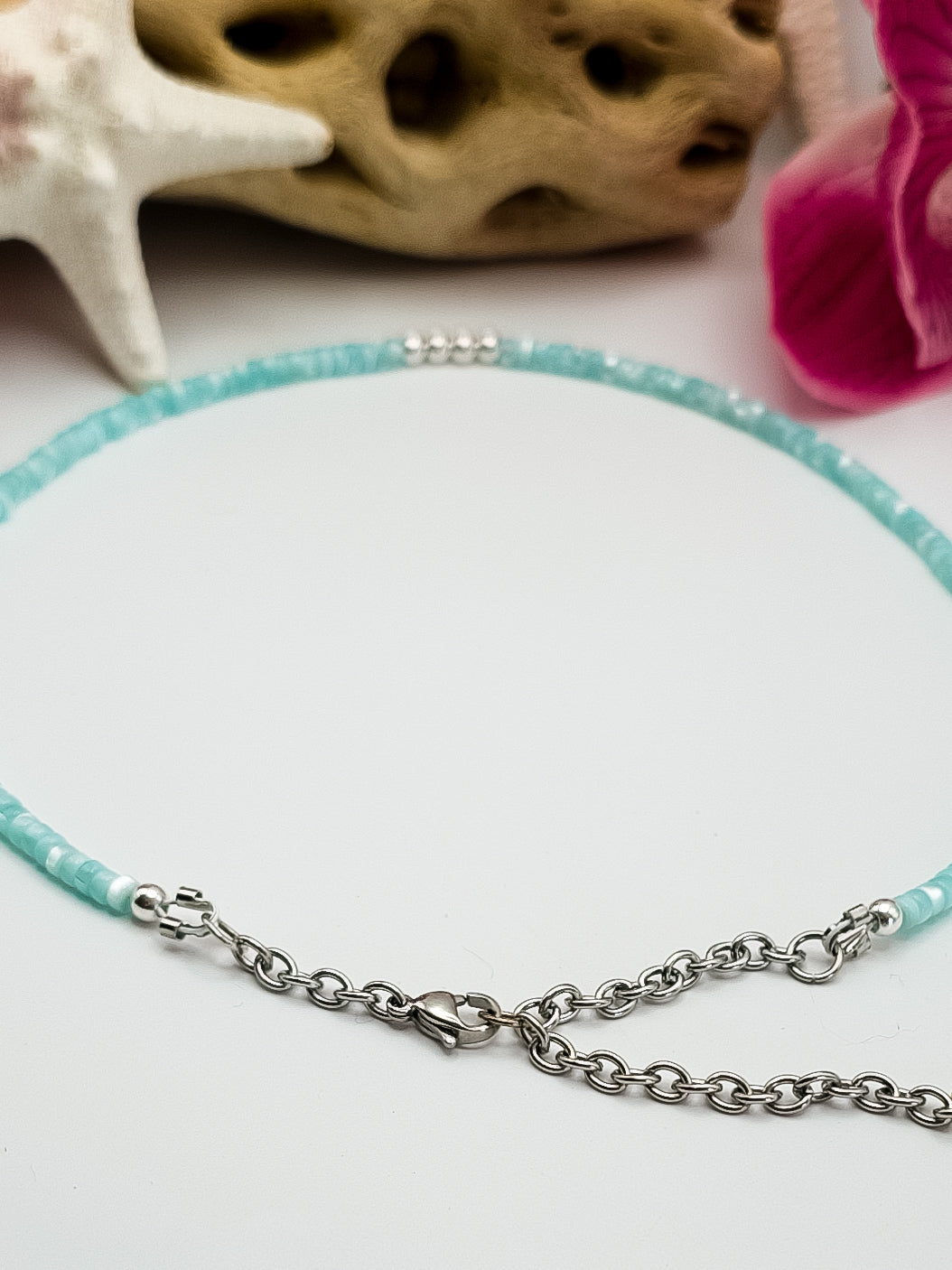 Turquoise Mother of pearl and silver beaded choker necklace with stainless steel extender chain and stainless steel lobster claw clasp laying on a flat white surface with flowers, driftwood, and starfish in the background for staging. 