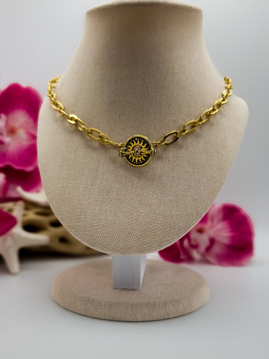 A gold-toned oval link chain necklace is displayed on a light brown jewelry bust. The necklace features a central, round toggle clasp as a pendant with a stylized sun design in gold and black. The sun has radiating lines and a central circle with smaller details.