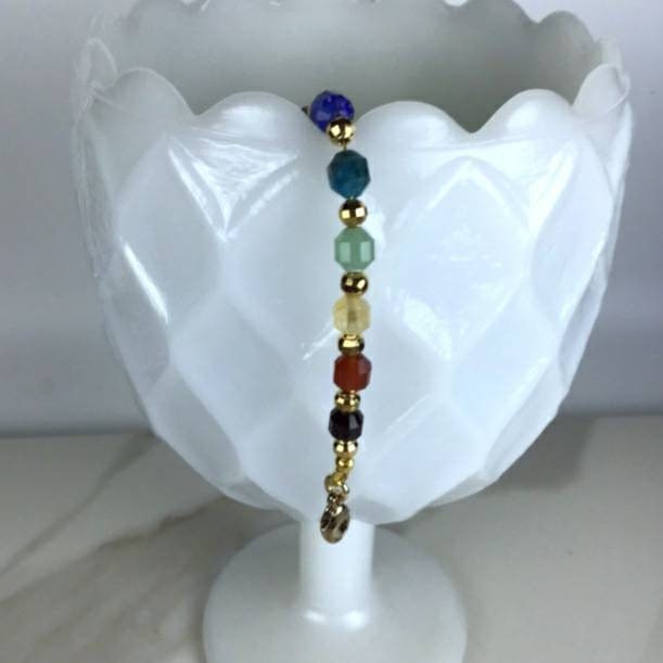 A chakra bracelet with a gold-filled chain features a sequence of gemstone beads in clear, purple, blue, light blue, green, yellow, orange, and red, representing the chakras. The bracelet hangs from the side of a white milk glass vase. 