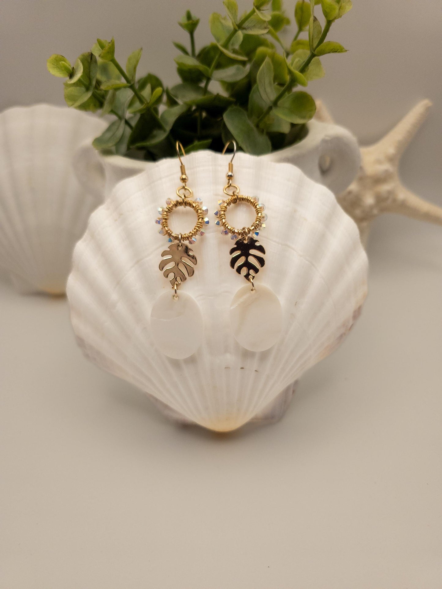 Gold-toned monstera leaf earrings adorned with shimmering Austrian crystals and luminous white Mother of Pearl shells. The earrings hang from a white shell against a textured vase filled with green leaves, creating a natural and elegant display. Seashells are visible in the blurred background.