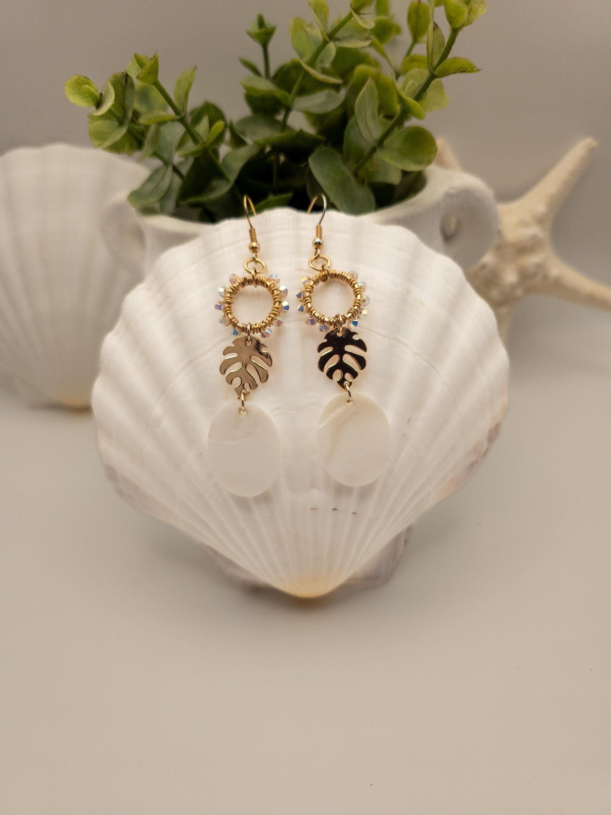 Gold-toned monstera leaf earrings adorned with shimmering Austrian crystals and luminous white Mother of Pearl shells. The earrings hang from a white shell against a textured vase filled with green leaves, creating a natural and elegant display. Seashells are visible in the blurred background.