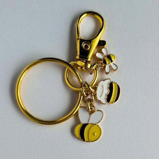 A gold-toned keyring features three enameled charms: a detailed honeybee, a small honey pot with dripping honey, and a tiny second honeybee. A swivel lobster claw hook is attached for versatile use as a keychain or bag charm.