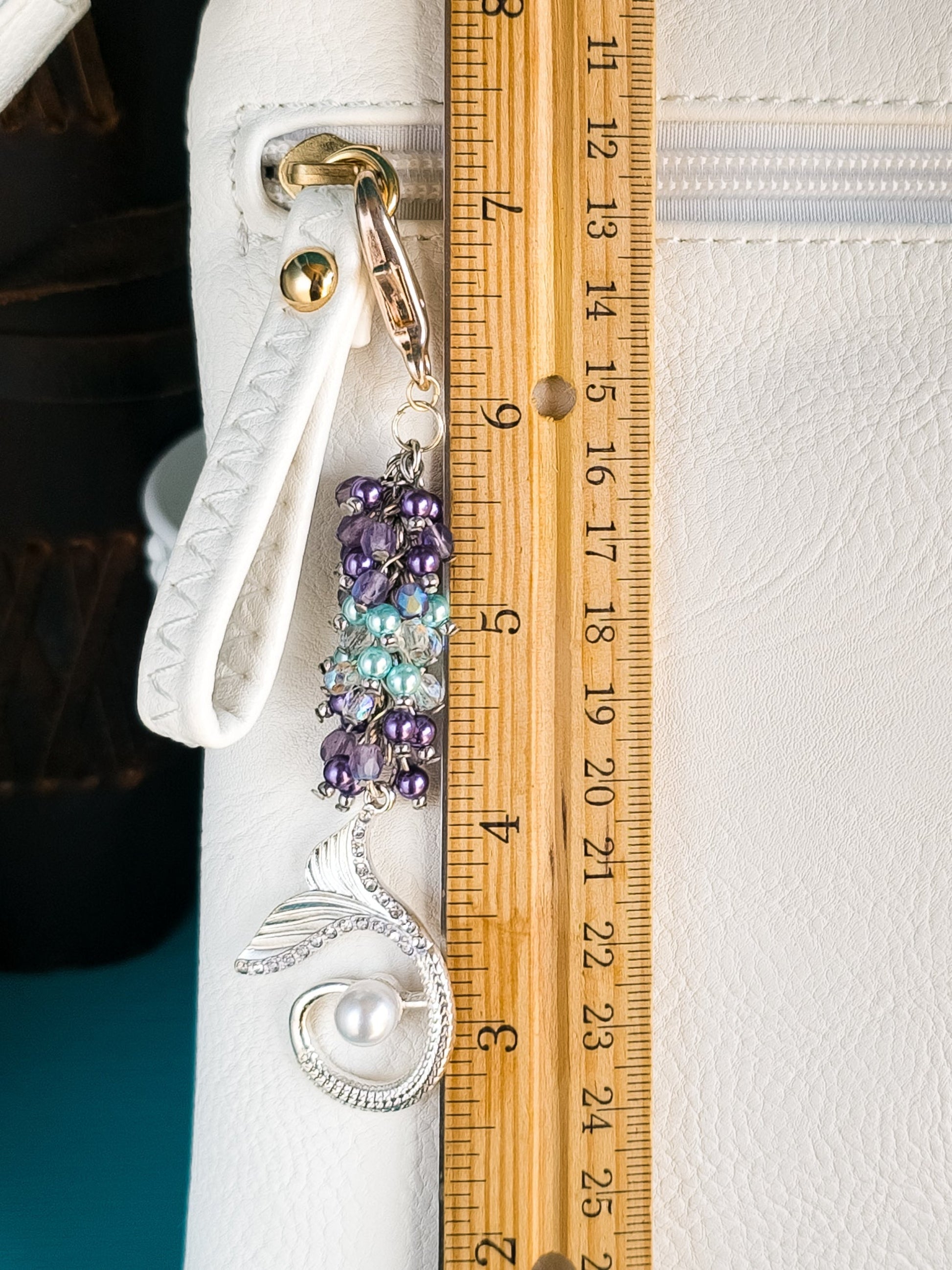 A mermaid bag charm, approximately 4.5 inches long as indicated by a wooden ruler, hangs from a white purse. The charm features purple and turquoise pearls, AB crystals, and a silver-plated mermaid tail.