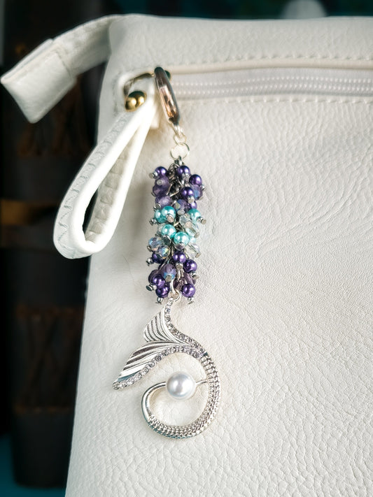 A mermaid bag charm, featuring purple and turquoise faux glass pearls, AB coated crystals, and a silver-plated mermaid tail with a white faux pearl, hangs from the handle of a white purse. The charm is attached with a large silver-plated lobster claw clasp and dangles from a stainless steel chain.