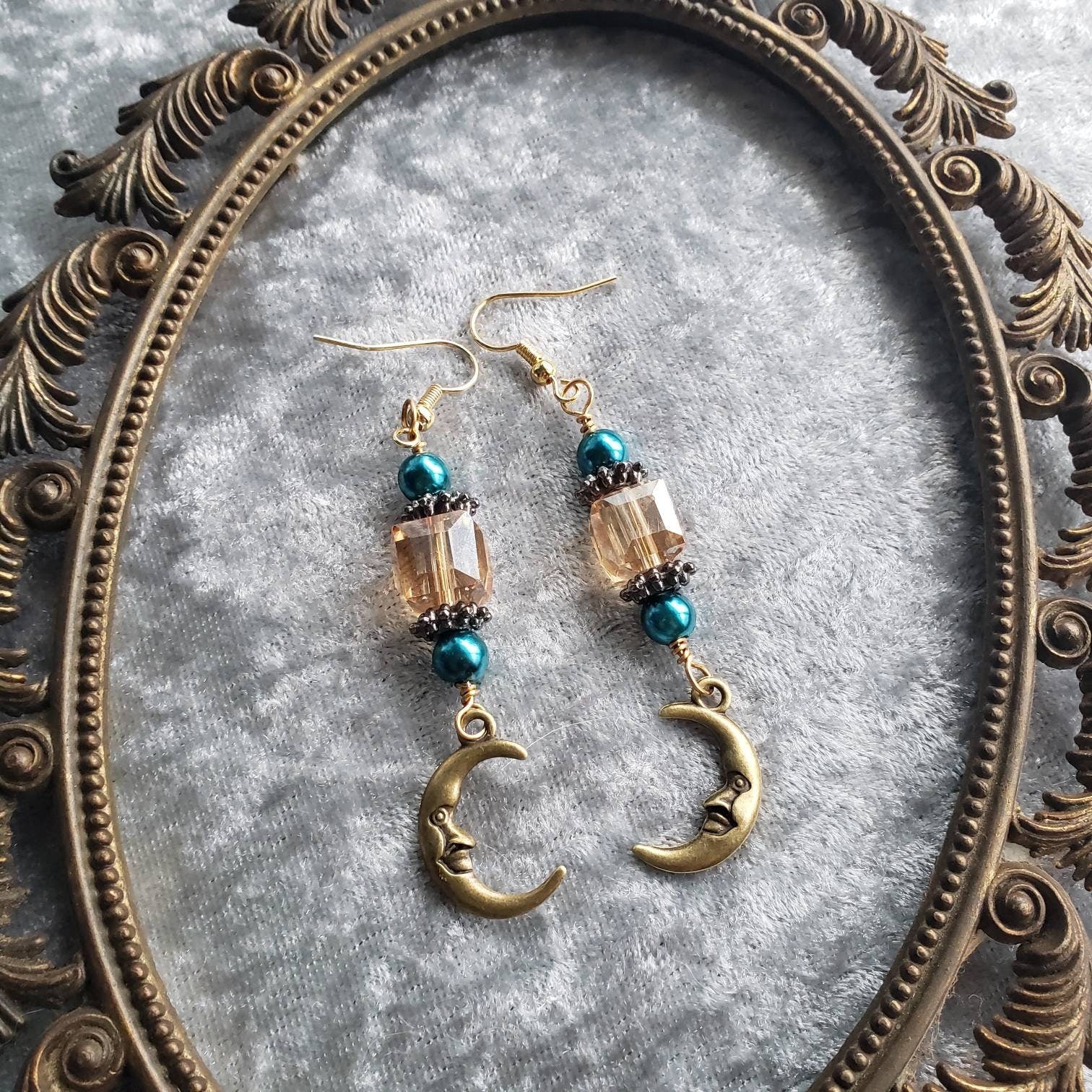 Dangle earrings composed of turquoise glass pearls, sparkling champagne crystals, and charming gold crescent moon charms with a small, endearing face. They are displayed on a soft silver velvet surface inside an ornate vintage gold frame.