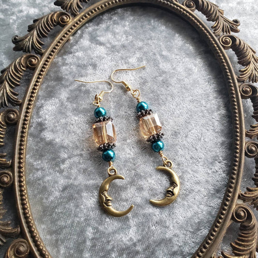 Dangle earrings composed of turquoise glass pearls, sparkling champagne crystals, and charming gold crescent moon charms with a small, endearing face. They are displayed on a soft silver velvet surface inside an ornate vintage gold frame.