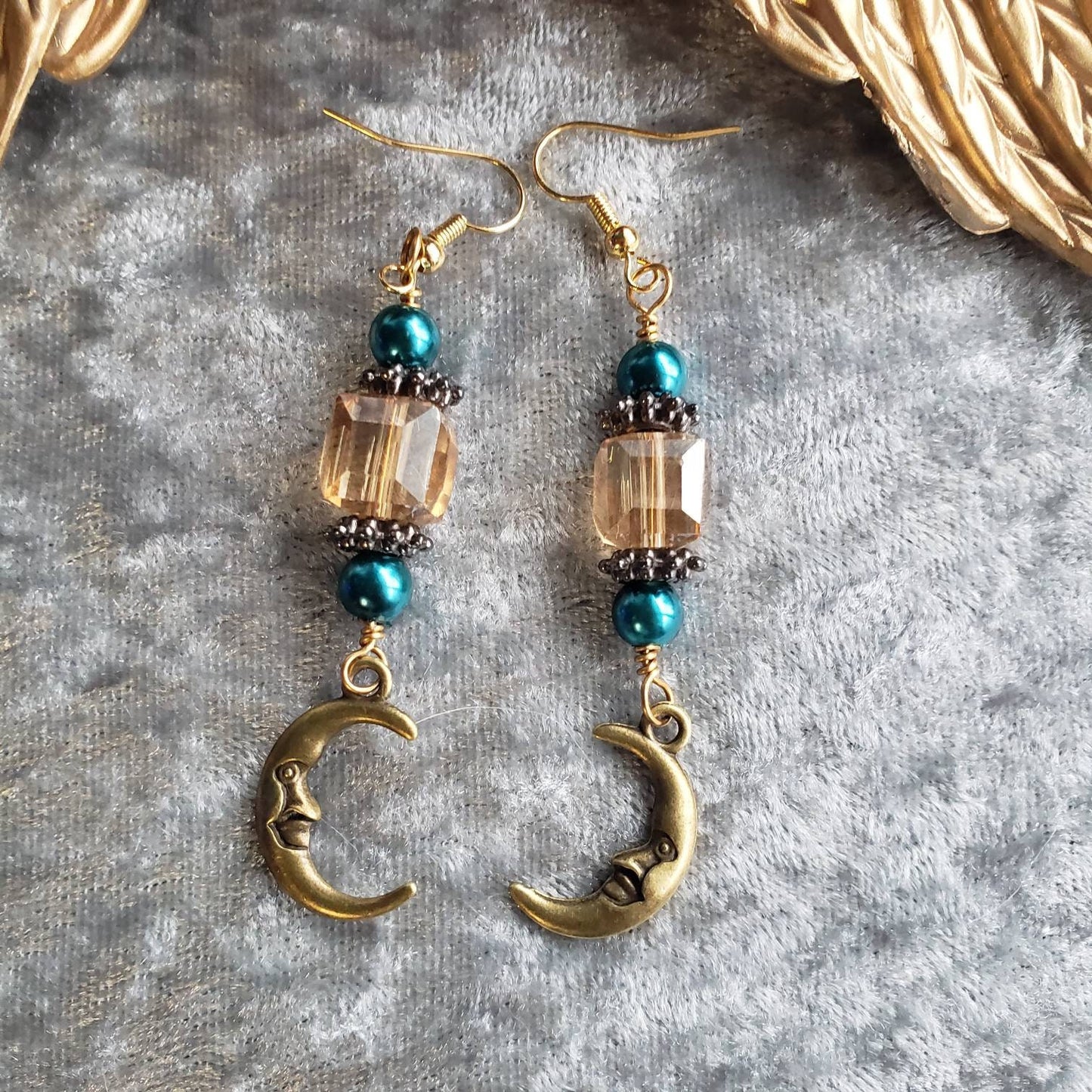 Whimsical dangle earrings with turquoise glass pearls, champagne crystals, and gold-tone crescent moon charms showcasing a cute, smiling face. The earrings are presented on a silver velvet backdrop within a decorative vintage gold frame.