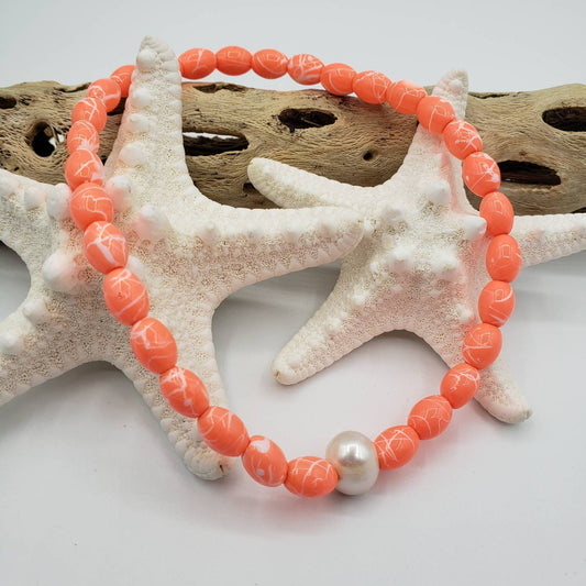 Stretchy orange beaded anklet featuring a single freshwater pearl as the focal point. The anklet rests across a collection of various starfish and weathered driftwood, suggesting a beachy setting.