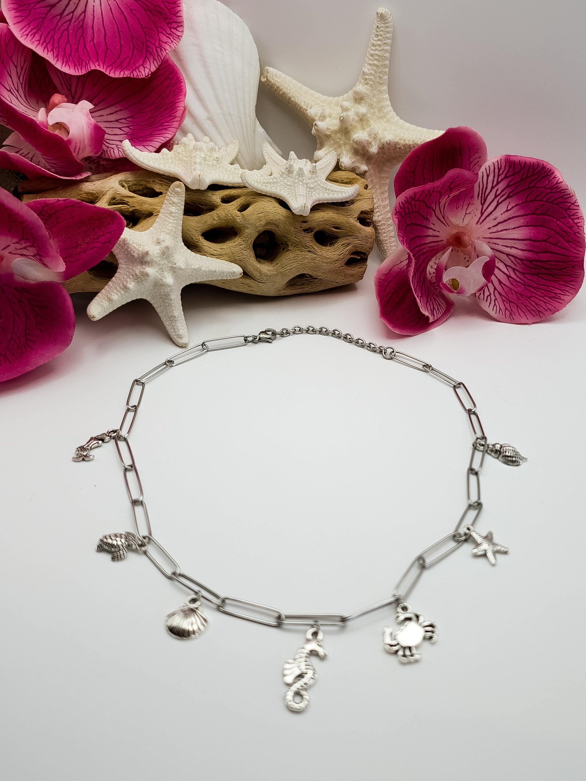 Stainless steel paperclip chain with beach charms in silver attached to the necklace. It is laying flat on a white surface. The charms are a mermaid, sea turtle, seashell, seahorse, crab, and starfish. Driftwood, fake starfish and flower in the background for staging. 