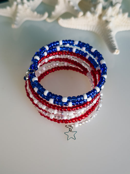 A multi-strand memory wire bracelet featuring red, white, and blue seed beads is the focus of the image. A silver wire star charm hangs from the bracelet. The background includes a slightly blurred white starfish.