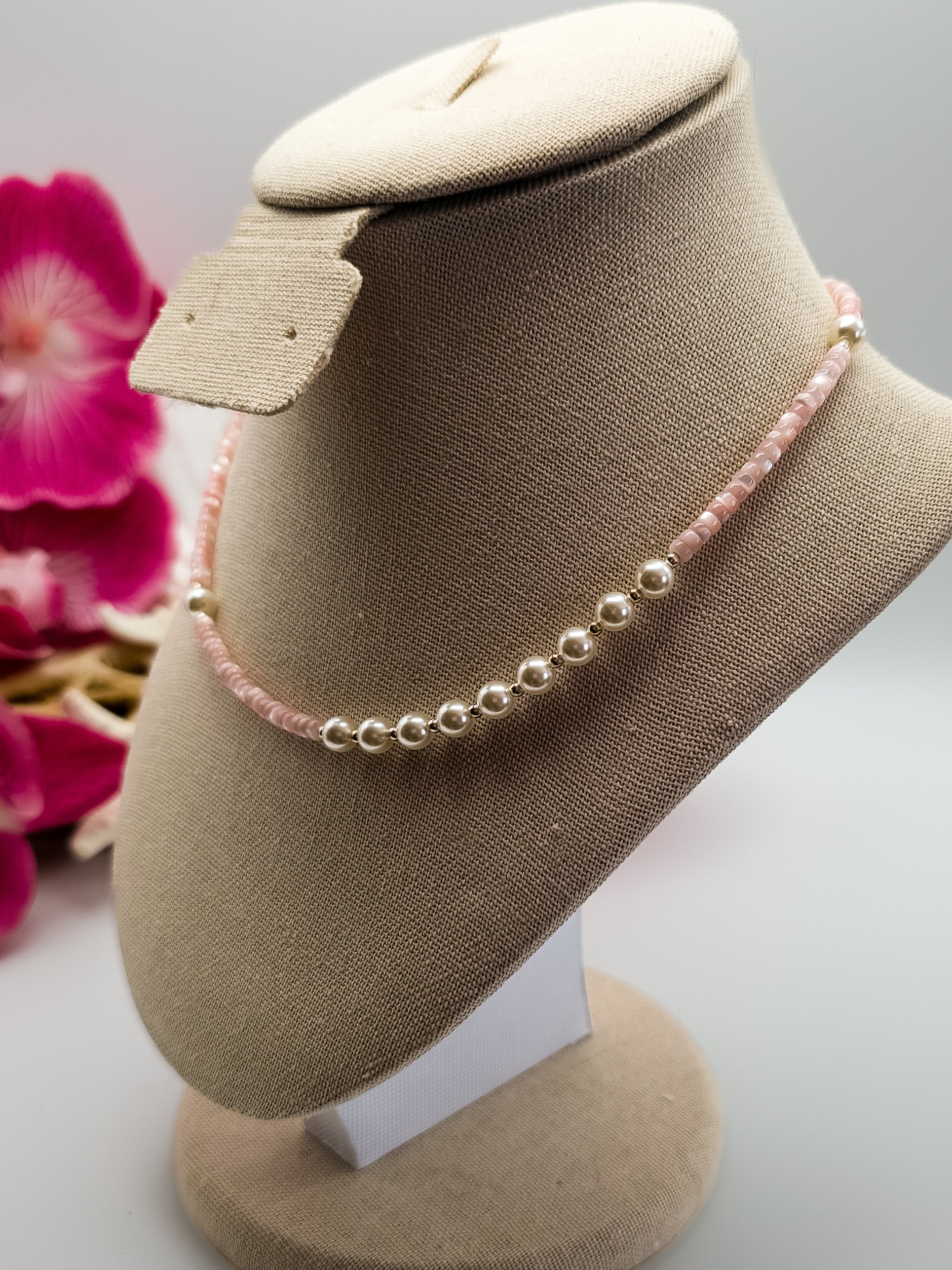 Light pink Mother of Pearl heishi beads with small silver plated beads and white glass pearls handmade choker laying on a tan neck display with flowers and driftwood in the background for staging. 