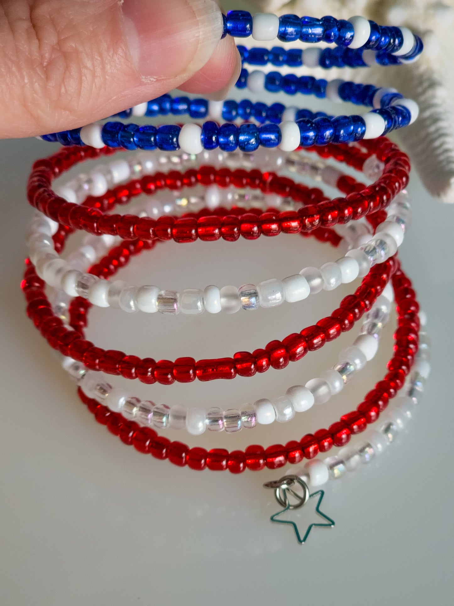 A detailed view of a memory wire bracelet featuring multiple strands of red, white, and blue seed beads. A silver outline of a five-pointed star charm hangs from one end. A hand is partially visible, holding the bracelet.