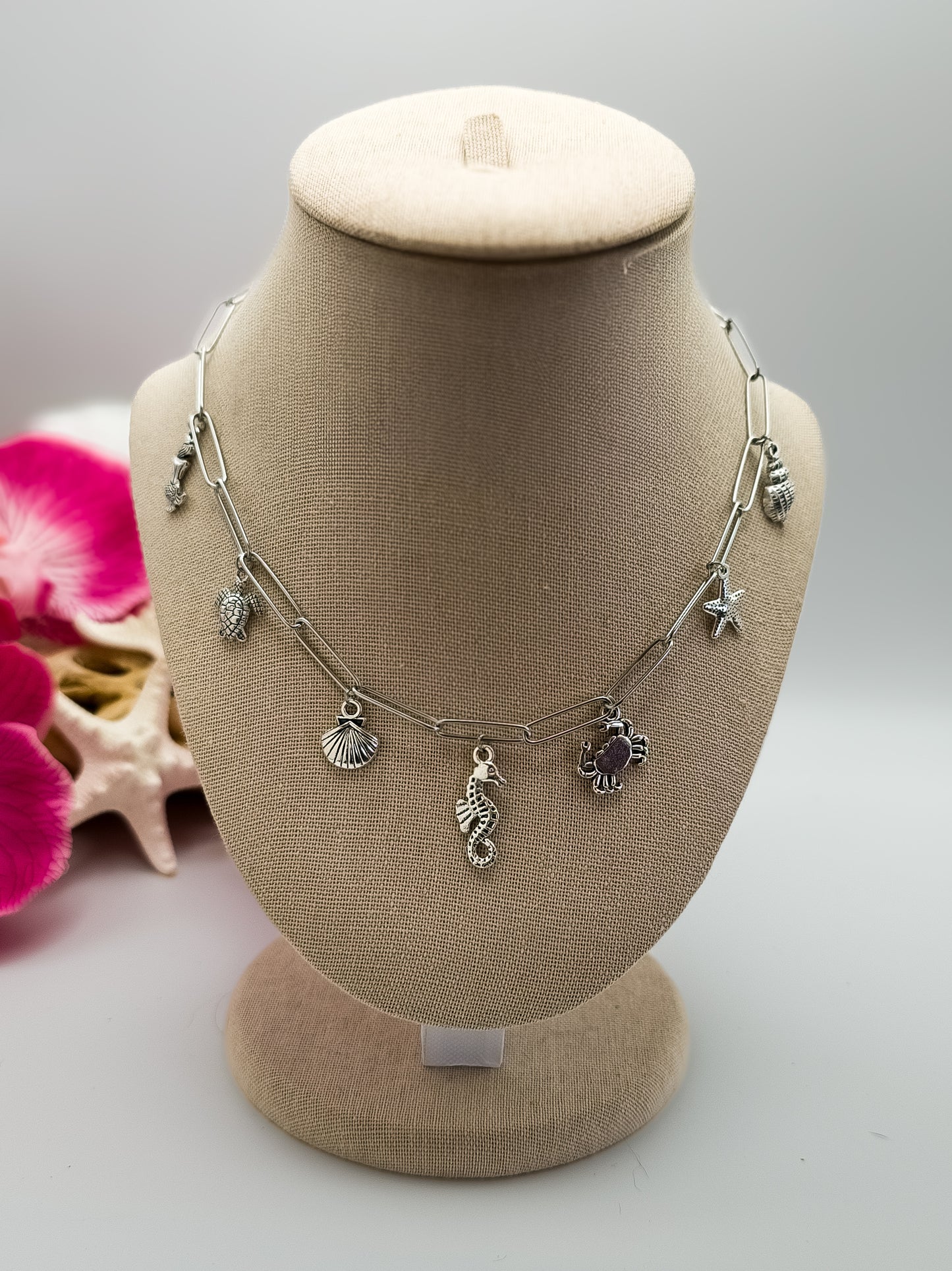 Stainless steel paperclip chain with beach charms in silver attached to the necklace. It is on a tan neck form. The charms are a mermaid, sea turtle, seashell, seahorse, crab, and starfish. Driftwood, fake starfish and flower in the background for staging. 