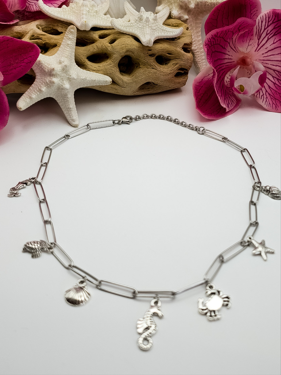 Stainless steel paperclip chain with beach charms in silver attached to the necklace. It is laying flat on a white surface. The charms are a mermaid, sea turtle, seashell, seahorse, crab, and starfish. Driftwood, fake starfish and flower in the background for staging. 