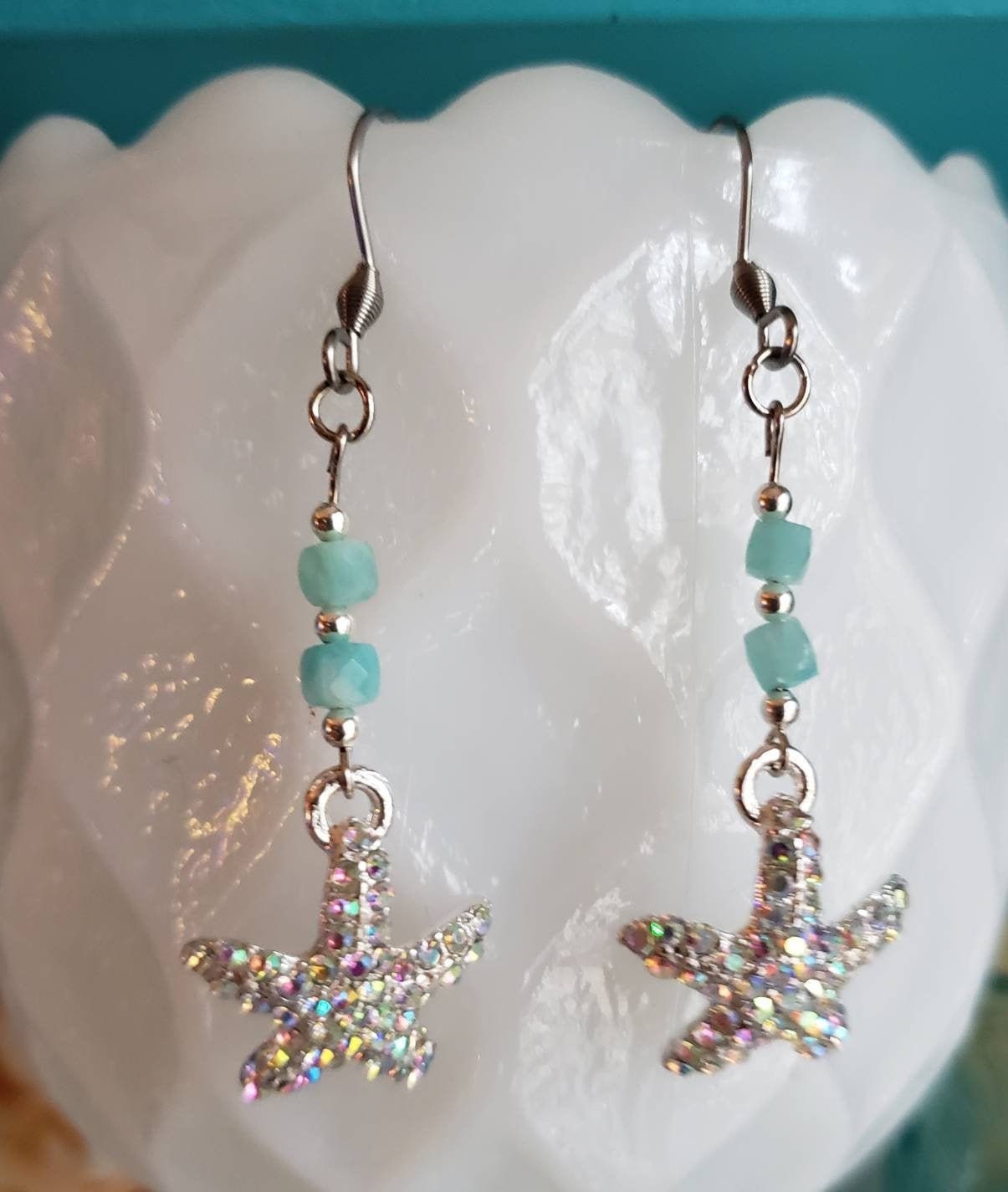 Silver starfish earrings adorned with sparkly rhinestones and blue-green amazonite beads dangle from a white milk glass vase. The vase provides a clean and elegant backdrop.