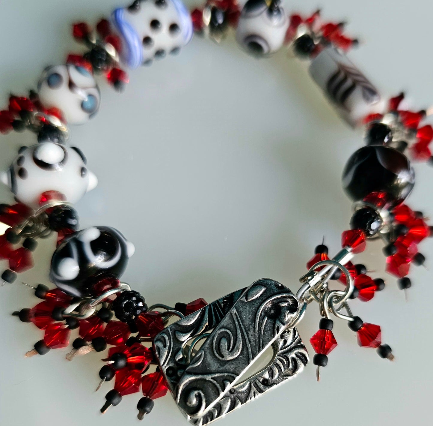 Handcrafted black and white lampwork beaded bracelet with vibrant red crystal accents. A detailed view showcases the antique silver rectangular toggle clasp with a subtle swirl design. The bracelet rests on a clean white surface.