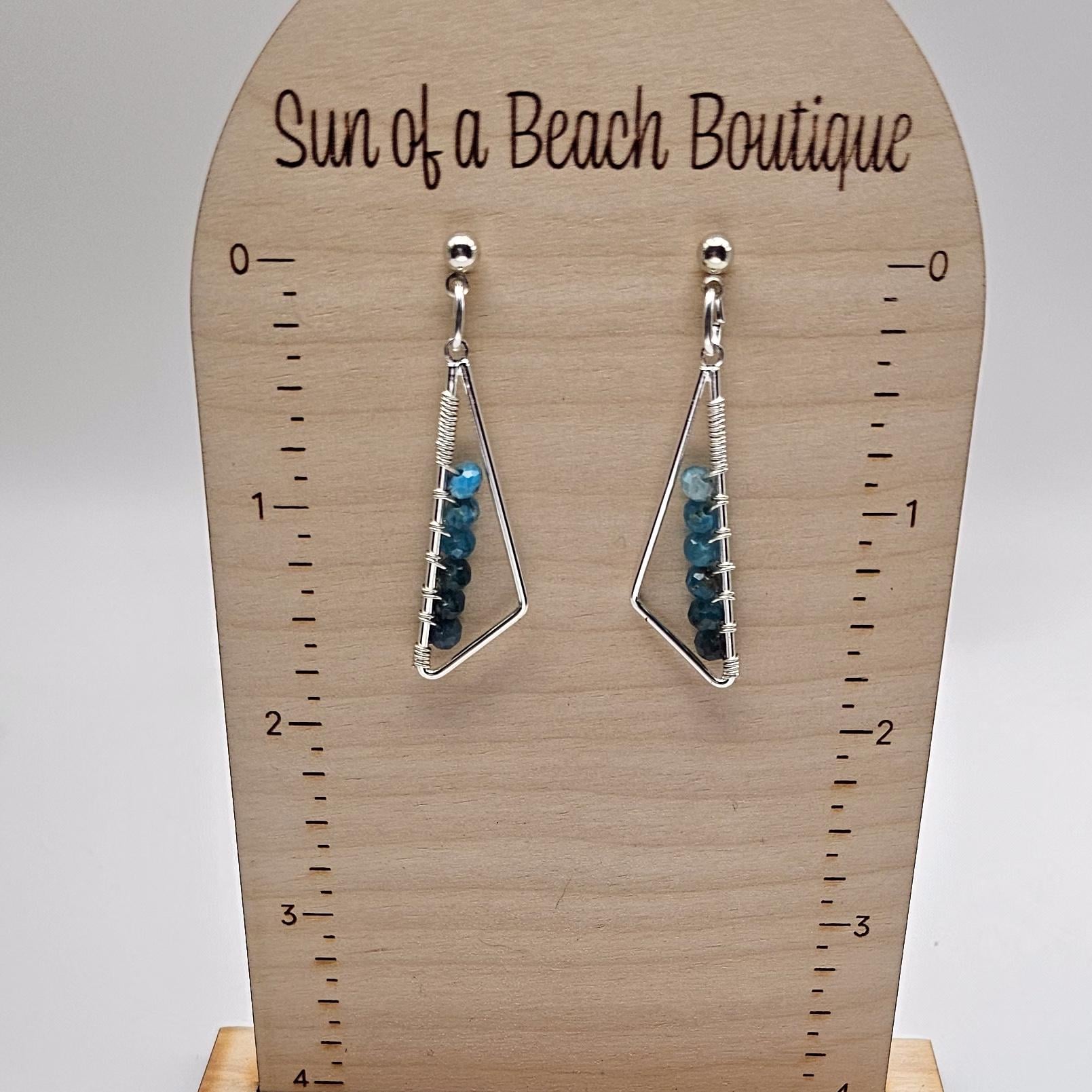 Silver triangle earrings with blue gemstone beads hang from an earring holder. The text "Sun of a beach boutique" is visible at the top of the holder. 