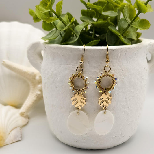 Elegant gold-plated monstera leaf earrings, each featuring sparkling Austrian crystals and a delicate white Mother of Pearl shell, dangle gracefully from a white rustic vase. Lush green foliage is arranged in the vase, and scattered seashells provide a subtle beachy backdrop.