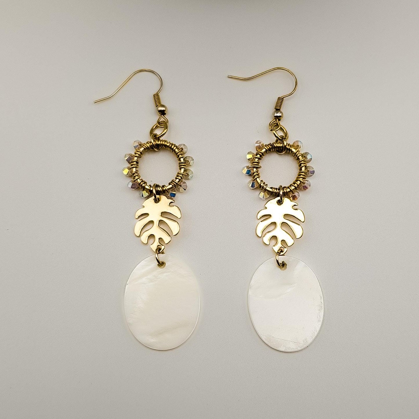 Close-up of gold-toned monstera leaf earrings adorned with shimmering Austrian crystals and luminous white Mother of Pearl shells. The earrings are laying on a flat white surface next to one another. 