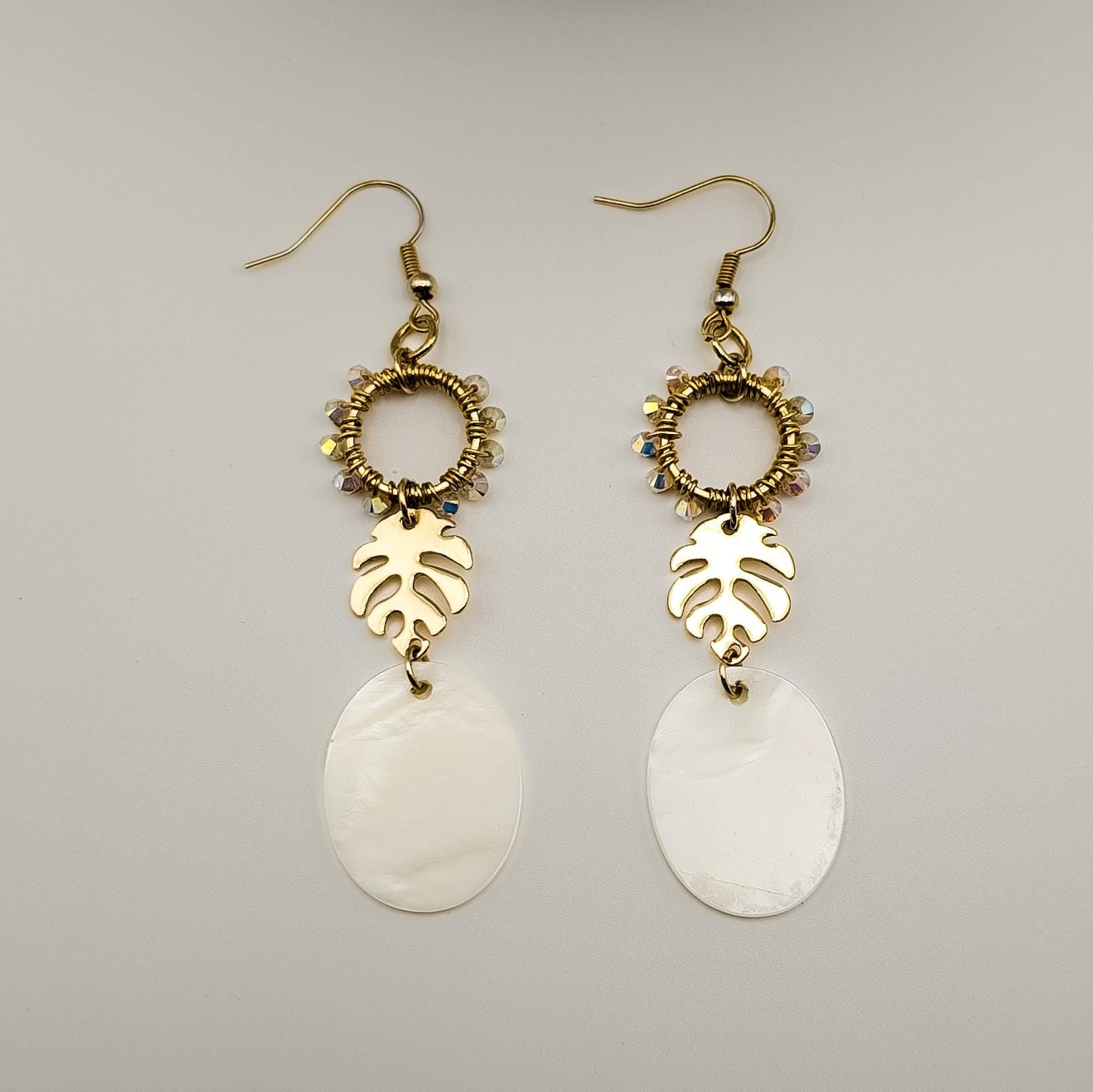 Close-up of gold-toned monstera leaf earrings adorned with shimmering Austrian crystals and luminous white Mother of Pearl shells. The earrings are laying on a flat white surface next to one another. 