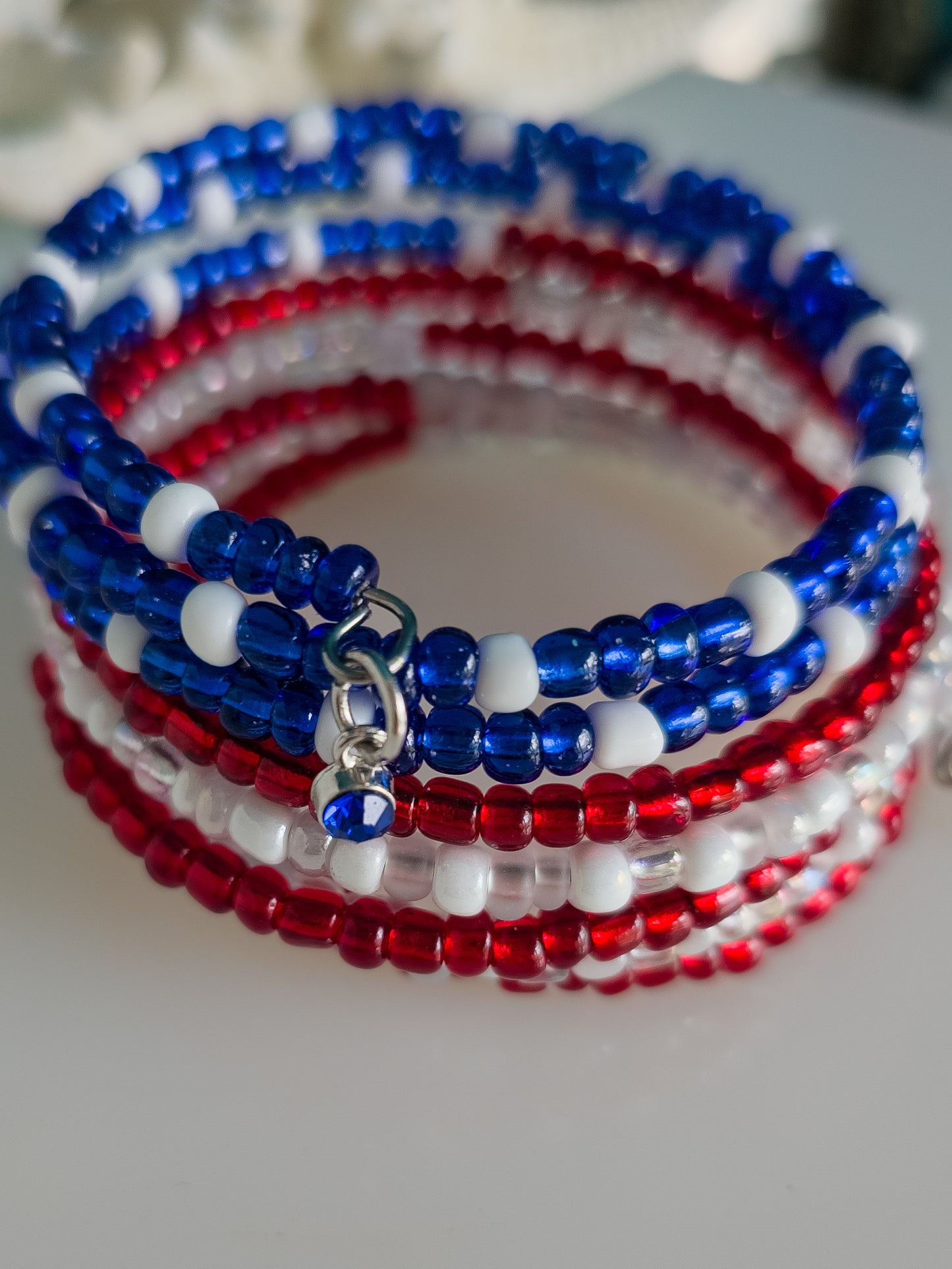 A detailed view of a memory wire bracelet featuring multiple strands of small red, white, and blue seed beads. A silver charm with a small blue bead dangles from the bracelet.