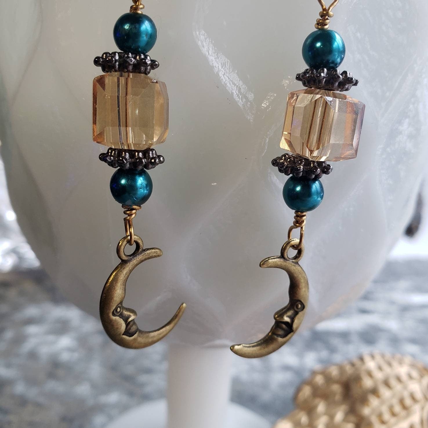 Whimsical dangle earrings with turquoise glass pearls, champagne crystals, and gold-tone crescent moon charms showcasing a cute, smiling face. The earrings are hanging from a white vase. 