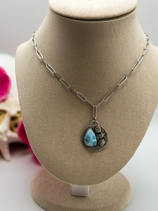Beach-themed paperclip silver necklace with a teardrop larimar stone and seashell and turtle accents displayed on a linen bust, with pink flowers in the background.