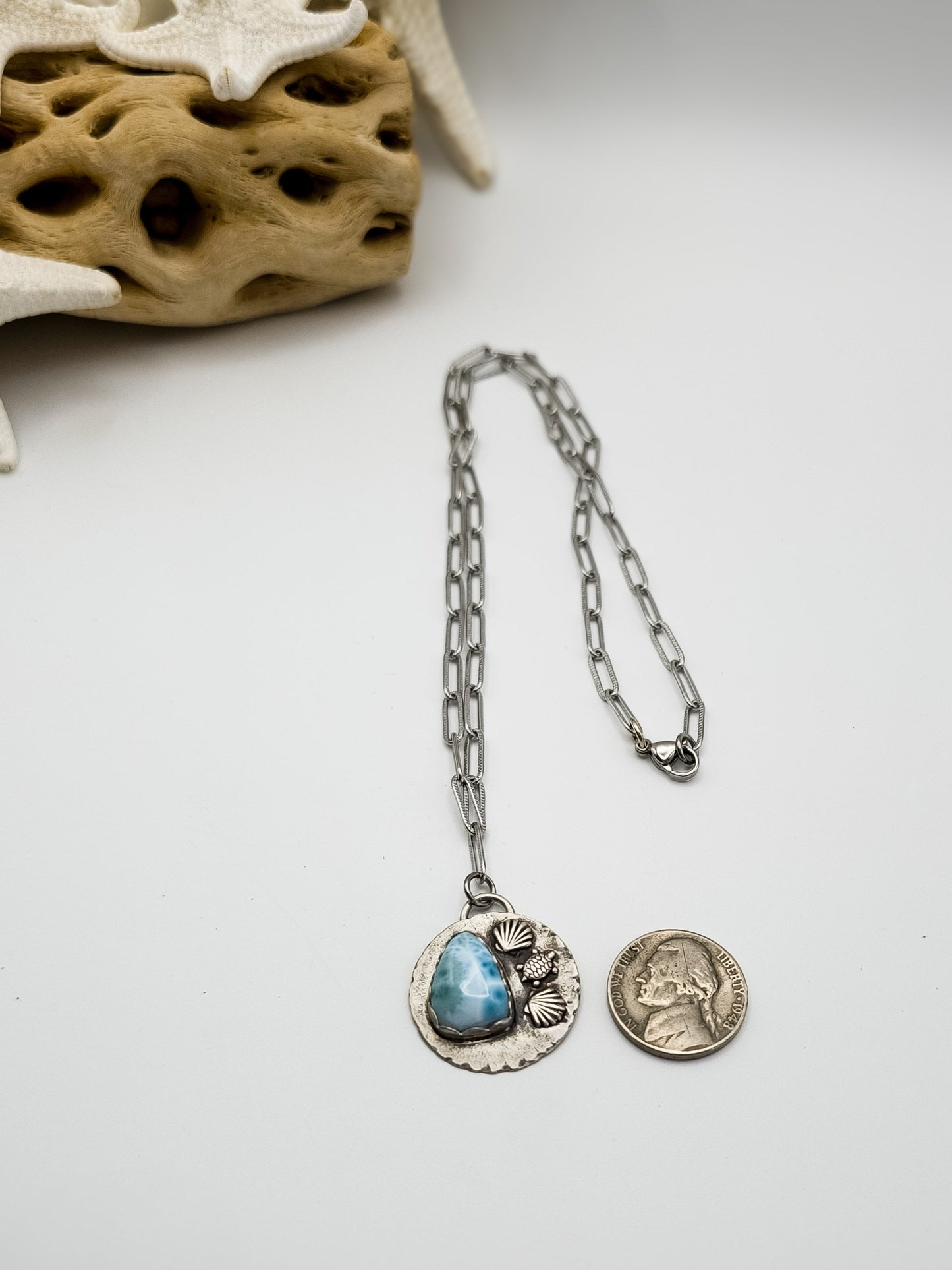 Beach-themed silver necklace with teardrop larimar gemstone pendant with shells and turtle accents on pendant with nickel next to pendant for comparison and with driftwood in the background.