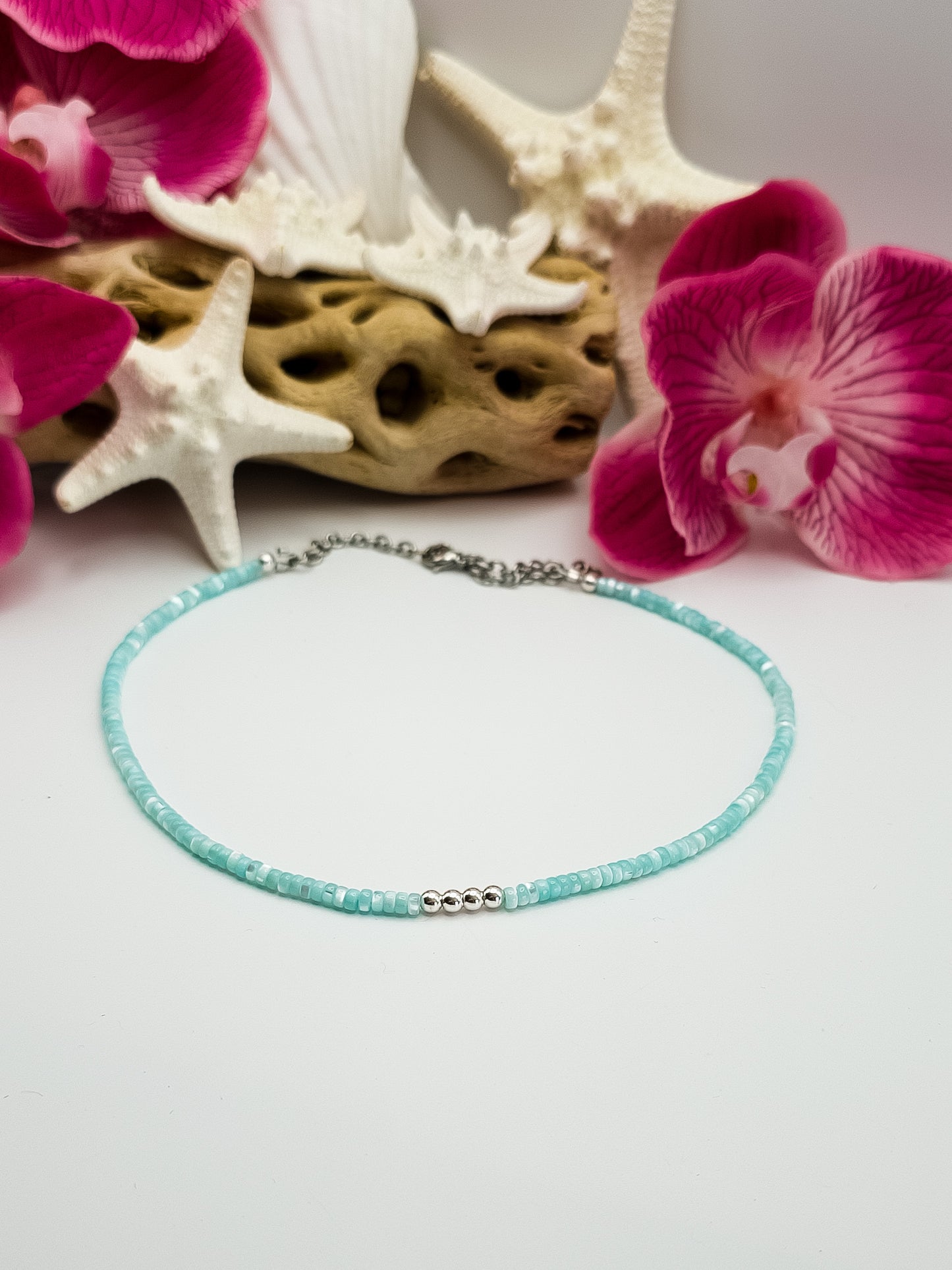 Turquoise Mother of pearl and silver beaded choker necklace with stainless steel extender chain and stainless steel lobster claw clasp laying on a flat white surface with flowers, driftwood, and starfish in the background for staging. 